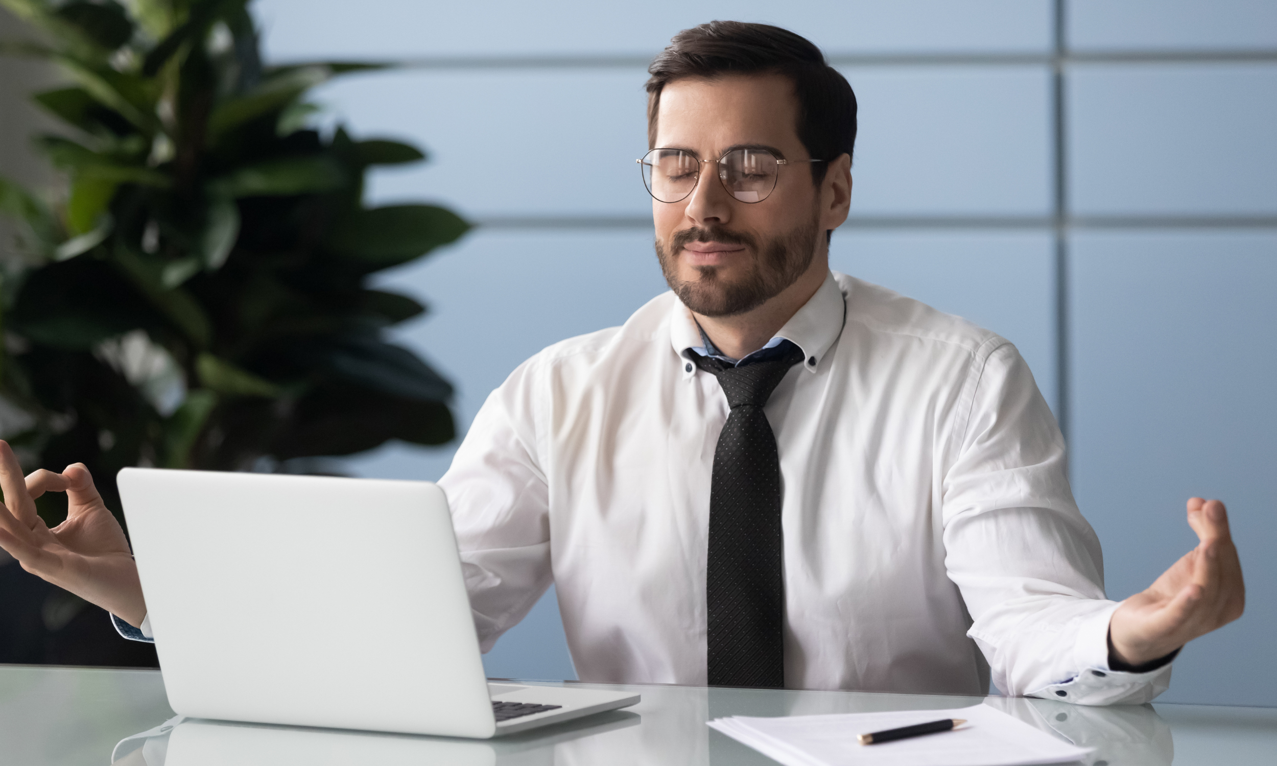 A man in a shirt and tie sits at a desk with crossed hands, laptop open, practicing mindfulness at work A man in a shirt and tie sits at a desk with crossed hands, laptop open, practicing mindfulness at work