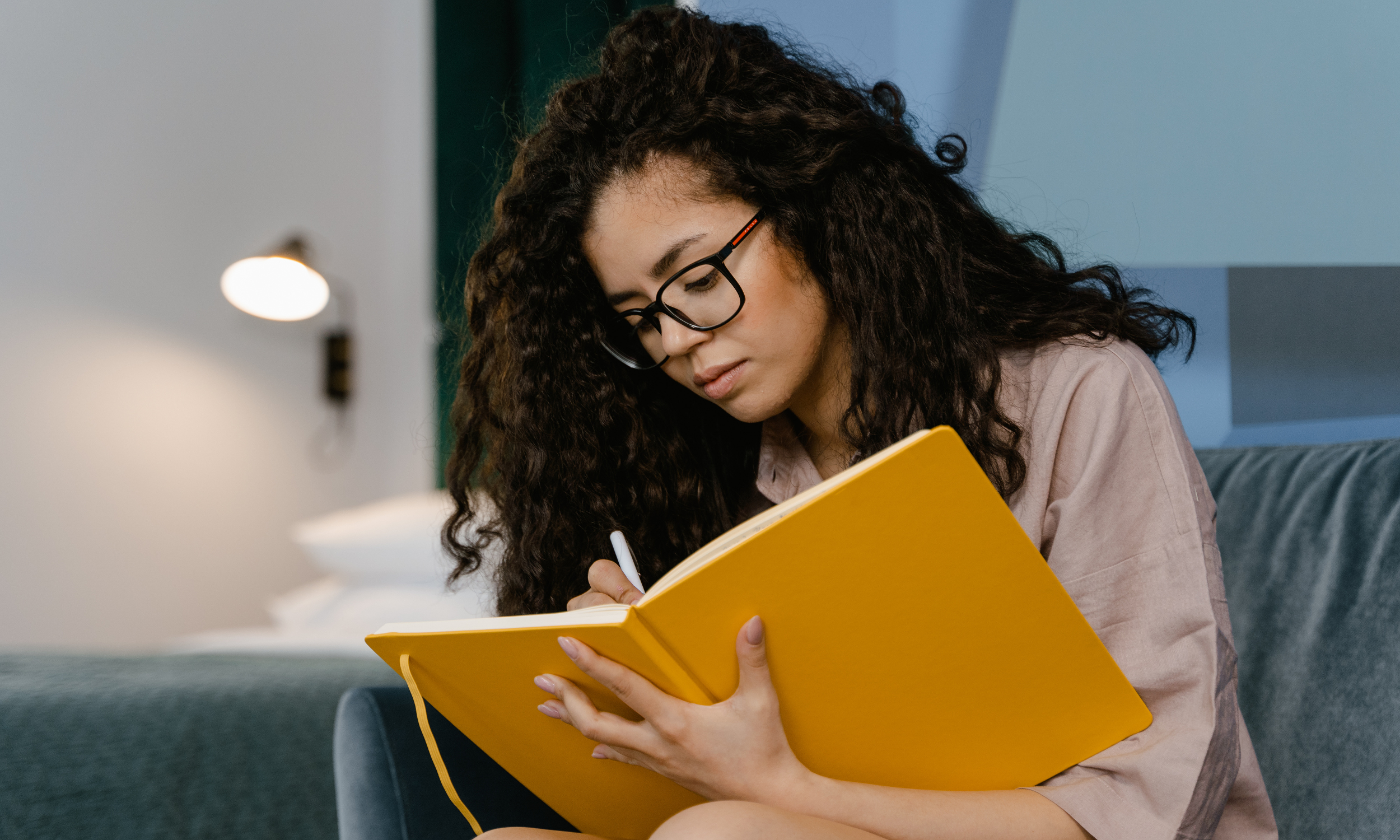 A woman wearing glasses is seated on a couch, engaged in journaling.